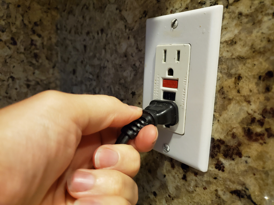 Close-up, personal perspective image of human hand of a man plugging an electrical cord into a Ground Fault Circuit Interrupter (GFCI) electrical outlet on the wall in a domestic room